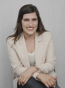 Smiling woman in a beige blazer seated with arms crossed against a grey background