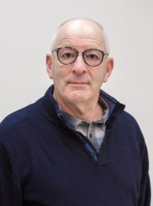 Portrait of an older man with glasses and grey hair in a navy sweater against a light background.