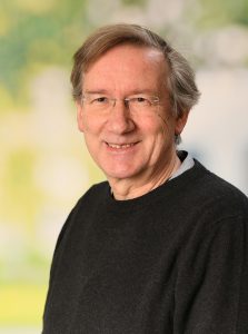 Portrait of a smiling older man with grey hair, rimless glasses and a dark sweater